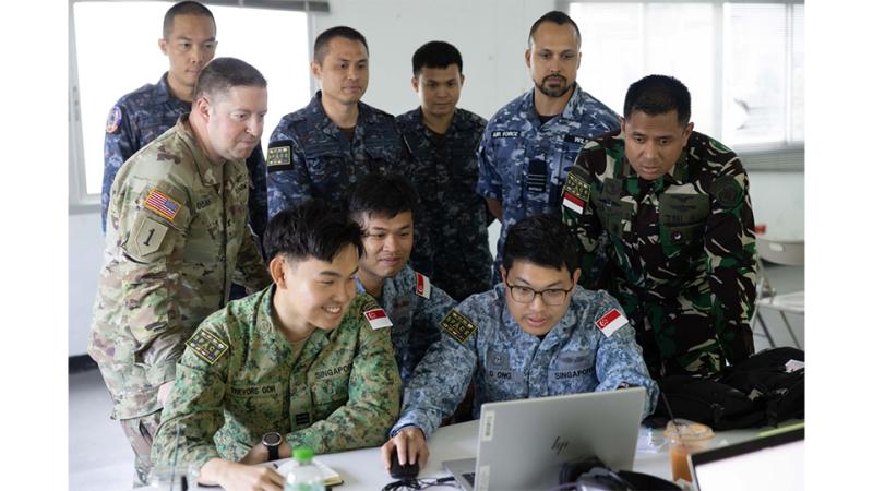 Several people in camouflage uniforms sit and stand around a laptop computer in a room.