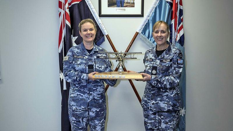 Two Air Force women hold a trophy shaped like an aircraft in front of two flags.