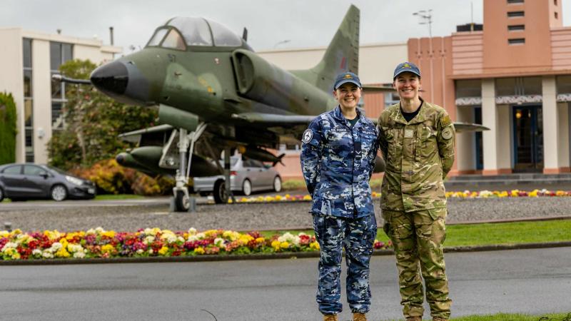 Two military members before an aircraft and brick buildings.