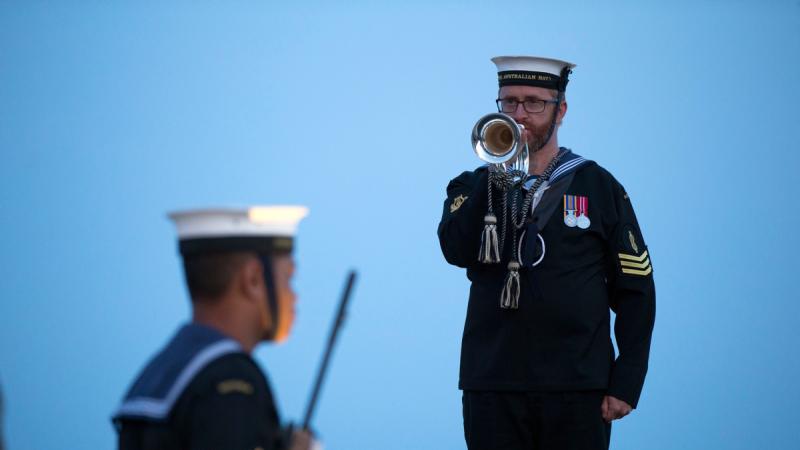 A Navy sailor plays a bugle at dawn, with another sailor blurred in the foreground against a blue sky.