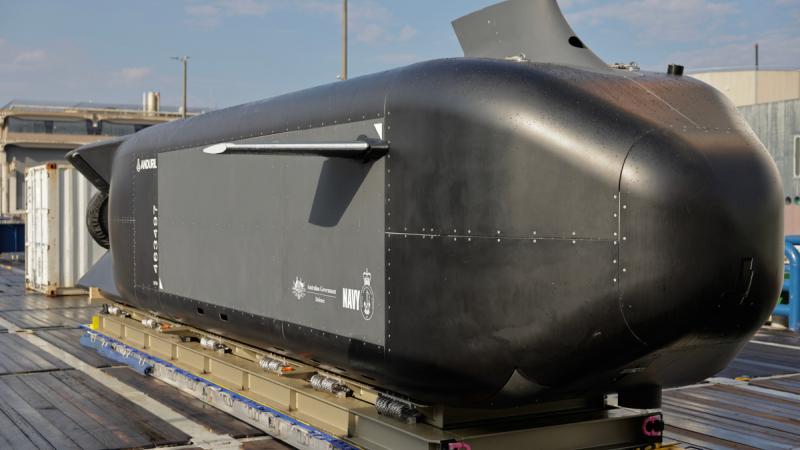 A black uncrewed underwater vehicle sits on a wooden surface in front of a shipping container.