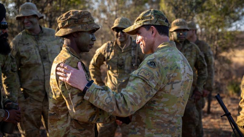 Two soldiers stand in the bush shaking hands, one with a hand on the other’s shoulder, with other soldiers visible in the background.