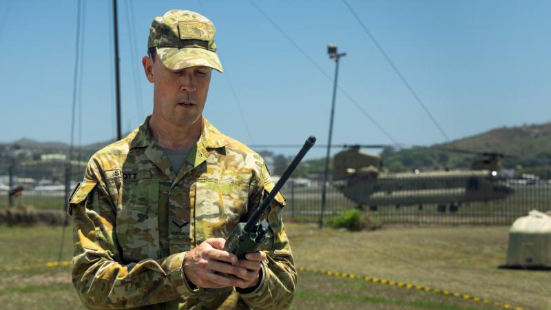 A uniformed man standing outdoors looking at a walkie-talkie in his hands.