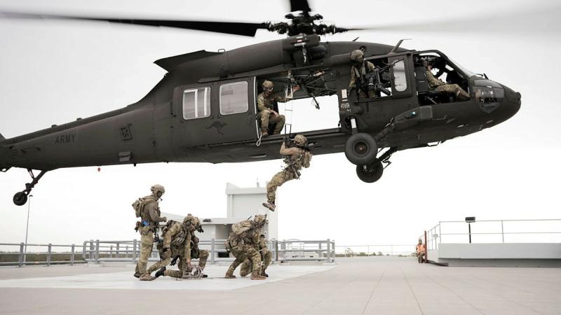 Soldiers crouch in a group beneath a helicopter with one soldier climbing on board.