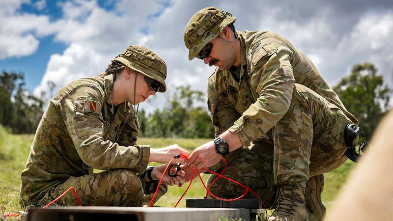 Two Army members crouch, handling wires in a grassy area.