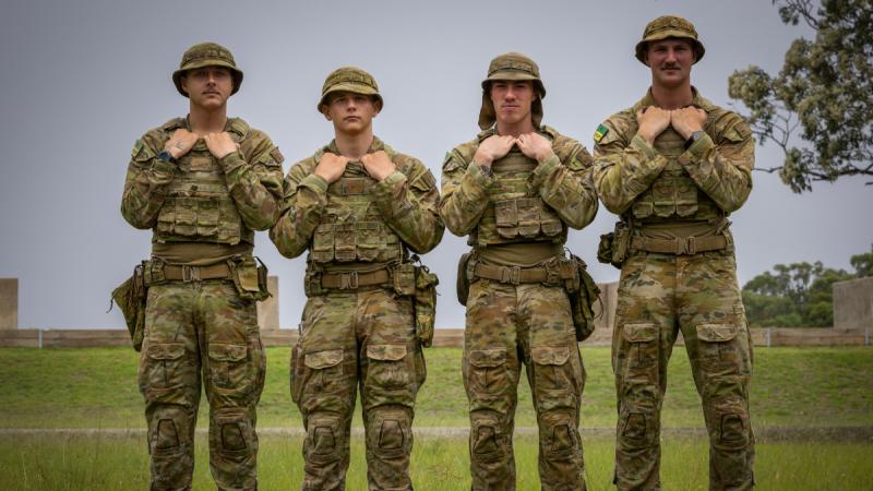 Four men in camouflage uniforms stand in a line in a field, gripping the collars of their uniforms.