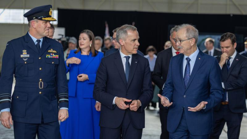 Three men, one in military uniform and two in suits, walk together while talking in a conference hall with other people in the background.