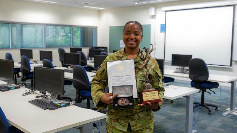A female Army member holds a certificate and award in a room full of desks and chairs.