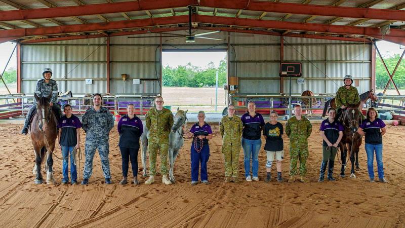 Defence personnel and civilians stand with horses in a large stable area.