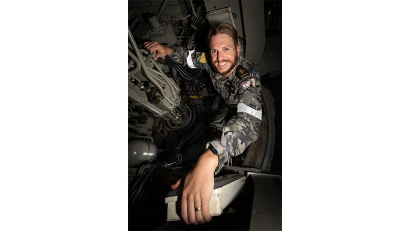 A man in camouflage uniform smiles while inside a confined mechanical space.