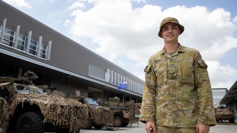 A man in uniform stands in a carpark in front of several camouflaged military vehicles.
