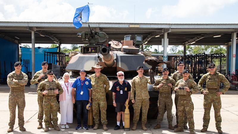 A group of soldiers and civilians standing together outdoors, with a tank behind them and one person wearing a glove with an eagle perched on their arm.