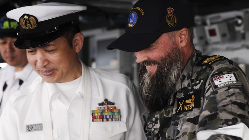 A Japanese and an Australian Navy member talk and look downward at the same area.