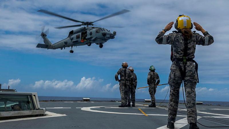 A Navy member directs a helicopter that is about to land on the flight deck of a ship.