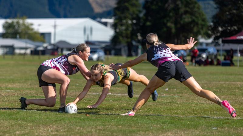 A player dives parallel to the ground to place the ball over the try line as two opposing players reach to touch her.
