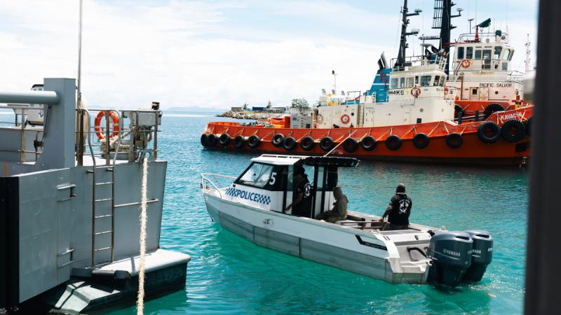 A police boat moves through clear blue water near a dock, with a larger tugboat moored nearby.