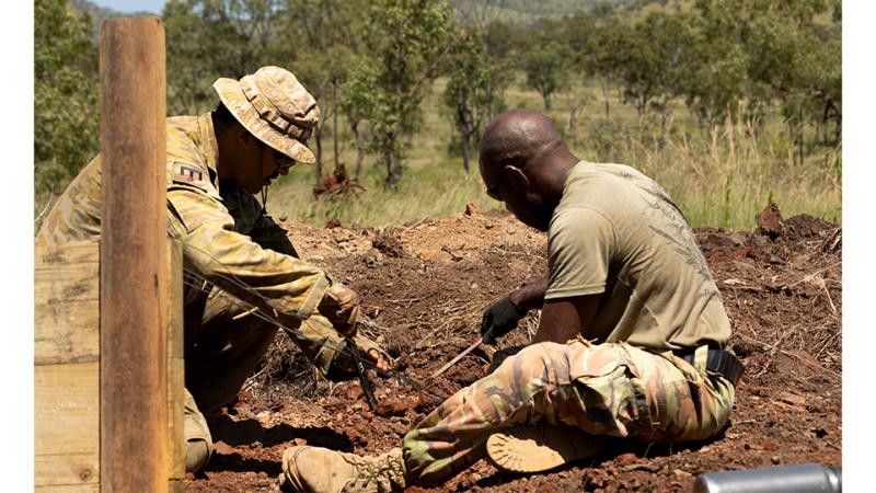 Two military personnel work together while seated on the ground outside.