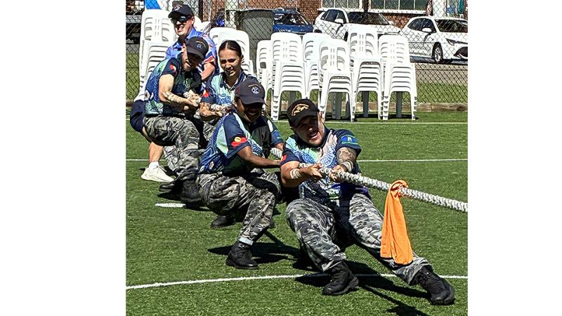 Five people lean back while pulling on a rope on an outdoor sports court, with white chairs and a fence behind them.