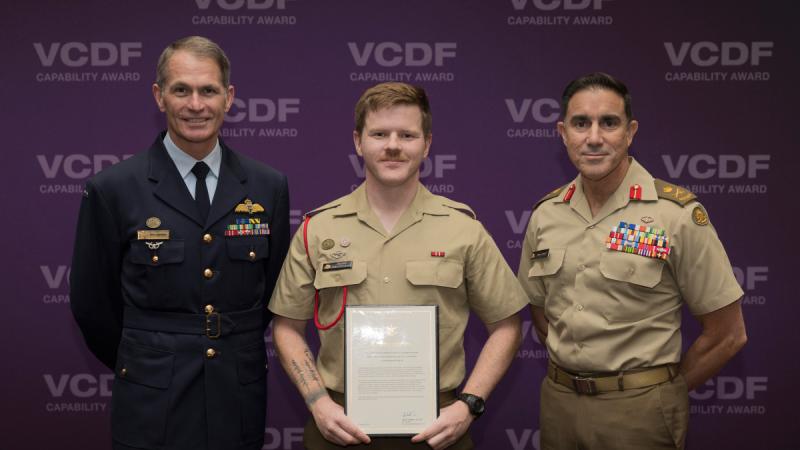Three men in formal uniform standing in front of a purple backdrop, the man in the centre holding a framed certificate.