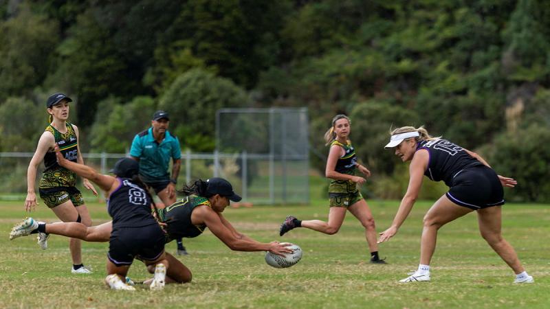 A female touch football player dives on the ball on a grassy field as others run towards her.