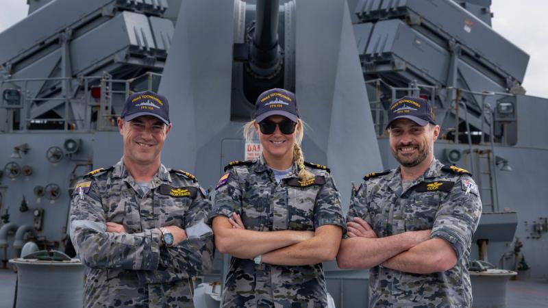 Three people in uniform stand on the forecastle of a ship beneath the 5-inch gun with their arms crossed in front of them.