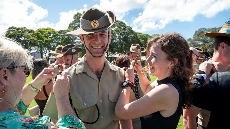 An Army member stands still while family add rank slides to his uniform.