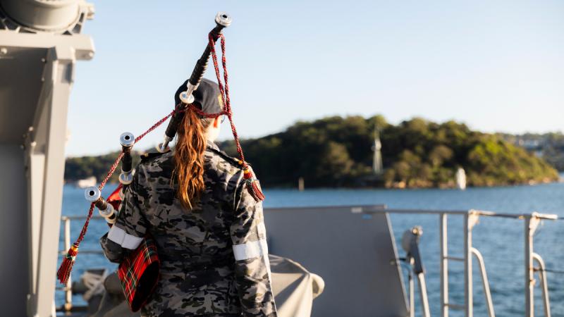 A woman in Navy camouflage holds bagpipes on the bow of a ship, facing a shoreline, seen from behind.