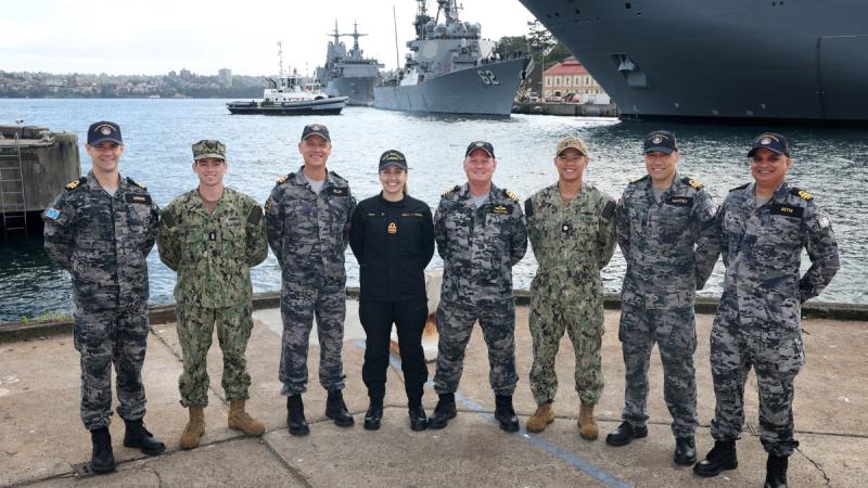 Several uniformed personnel stand along the harbour foreshore with docked warships visible across the water.