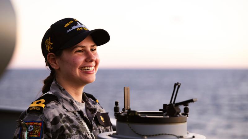 A woman in Navy uniform on a ship looking out to sea and smiling. 
