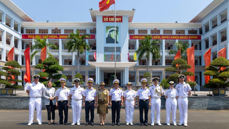 A group of people stand side by side in front of a large building decorated with red flags and Vietnamese writing.