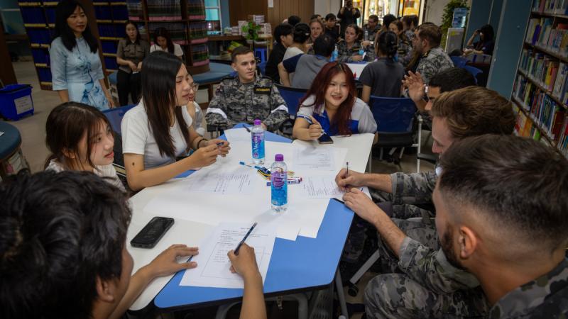 Men in camouflage uniforms and young women sit around a small desk in a classroom, talking.