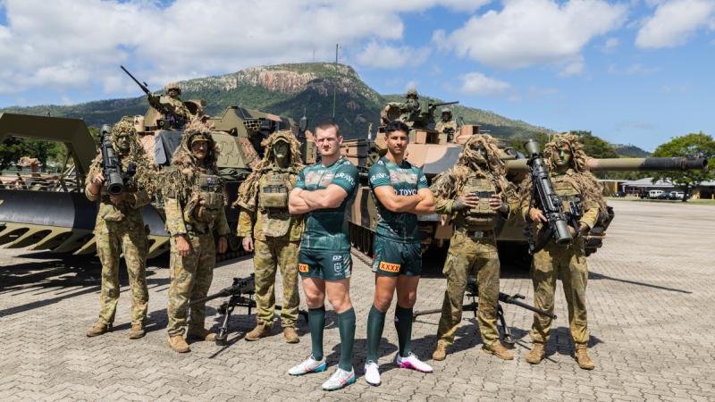 Two men in green sports uniforms flanked by soldiers in camouflage, with soldiers in tanks behind them and a mountain in the background.