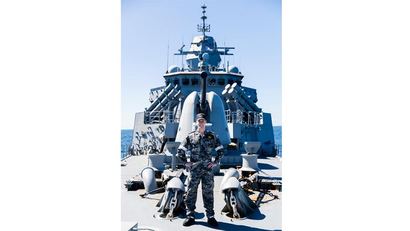 A man in camouflage uniform stands underneath a 5-inch gun on the deck of a ship.