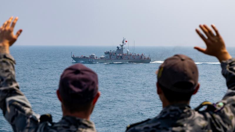 Two people in baseball caps and camouflage uniforms wave at a ship in the distance.