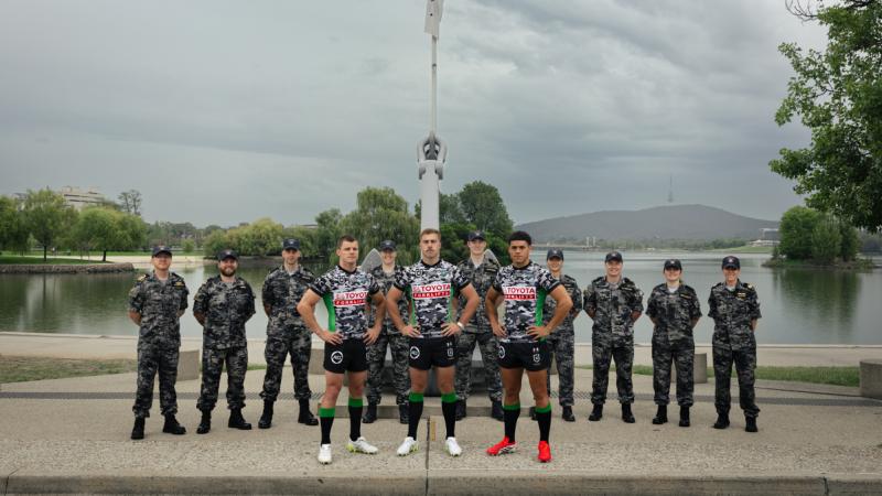 Navy personnel stand in a line at the base of a flag with three rugby league players in front of them.