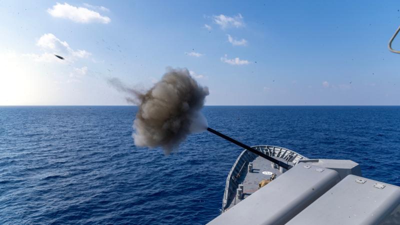 Grey smoke rises from a ship’s 5 inch gun as a projectile travels through the air over open sea.