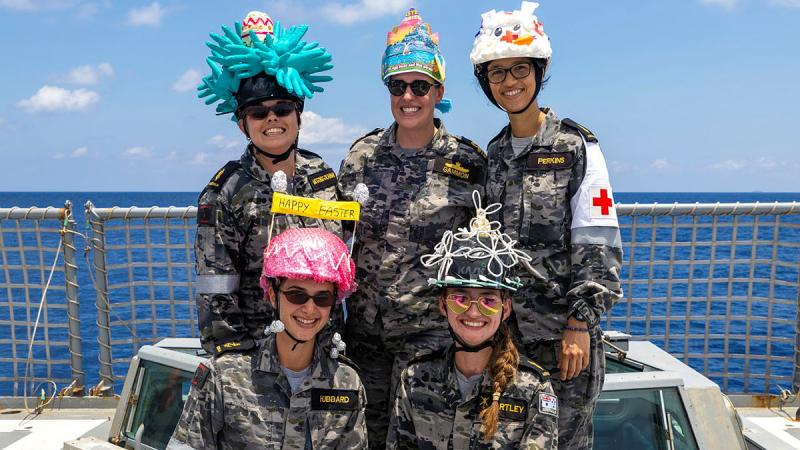 A group of sailors sit on the deck of a ship wearing Easter hats they have created.