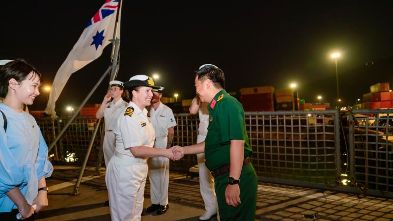 A woman in a white uniform shakes hands with a man in a forest green uniform on a ship’s deck, with the Australian White Ensign flying behind her.
