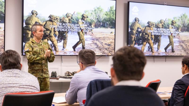 A uniformed man stands at the front of a classroom speaking to seated people, with large screens behind him.