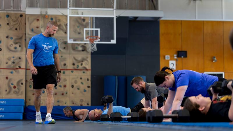 A man watches over a group of people doing push ups in a gym.