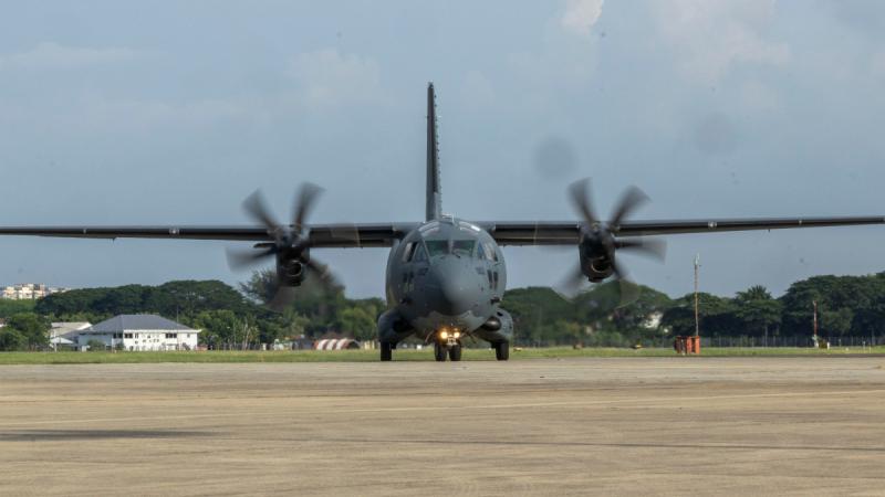 A grey military aircraft on a tarmac, its propellers rotating.