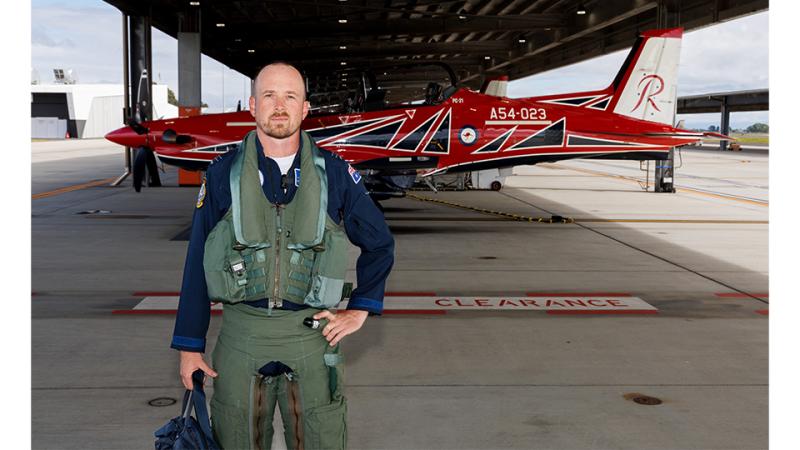 A man in flying uniform stands in front of an aircraft in a hangar.