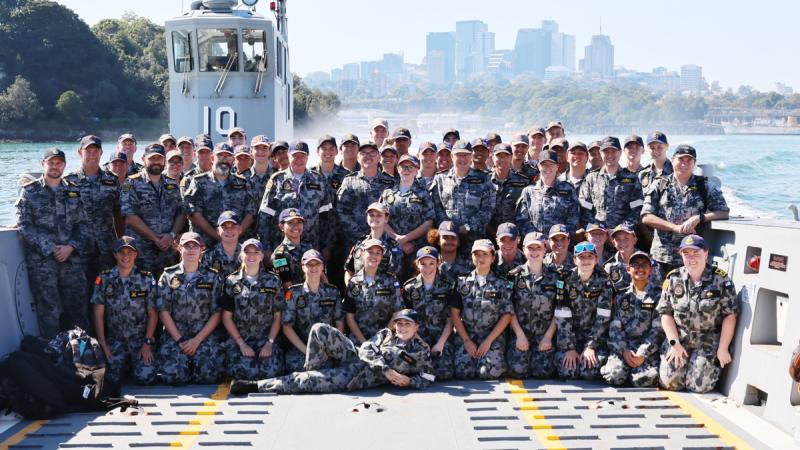 A large group of people gathered on the deck of a ship with a city skyline in the background.