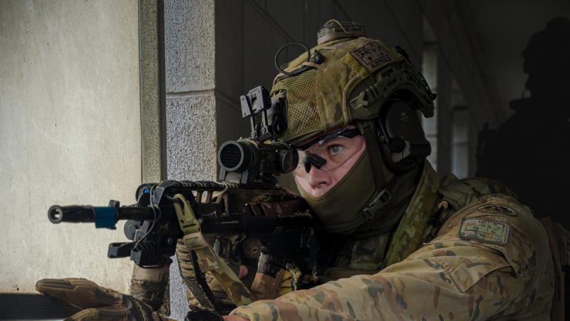 A man in full camouflage uniform aims a gun through a window.