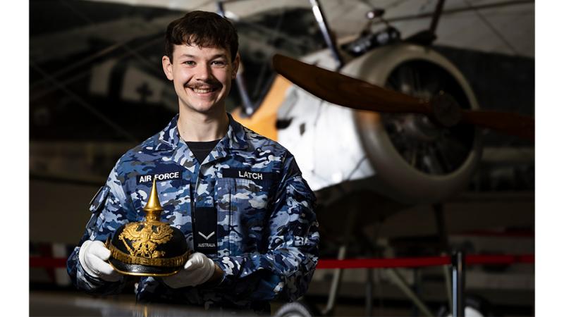 A military man stands before an aircraft with an elaborate helmet in hand. 