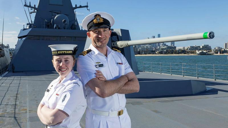 A shorter Navy member and a taller one stand back to back with their arms crossed, on the deck of a ship.