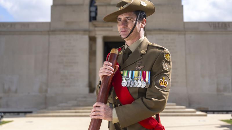 A man in Army uniform holding a didgeridoo in front of a memorial structure.