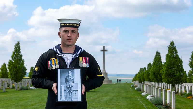A sailor stands in a war cemetery holding a photo of a relative.