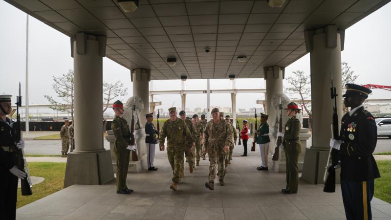 Military personnel walk underneath an archway guarded by armed military personnel.