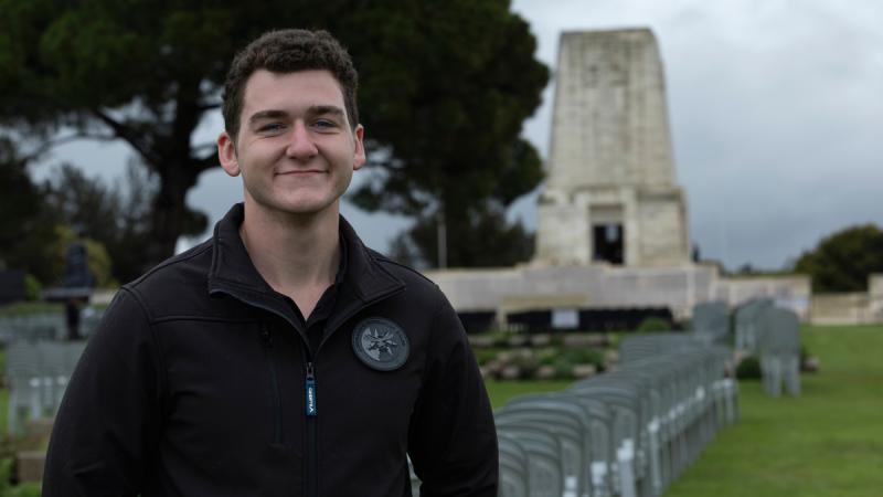 A man standing among headstones at a cemetery, with a large memorial and tree in the distance.